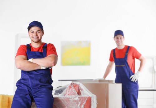 The image depicts two professional movers dressed in red shirts, blue overalls, and matching blue caps, working indoors in a residential setting, likely in a house in Richmond. The mover in the foreground is smiling confidently with his arms crossed, standing beside a large cardboard box partially wrapped in protective plastic, which is positioned on top of a wheeled trolley. The second mover, positioned slightly further back, stands with one hand resting on his hip and the other on the box, both appearing prepared to lift or move items. The background shows a bright, well-lit room with white walls and minimal decor, including a small abstract painting or print in yellow and white tones hanging on the wall. A portion of a yellow sofa or armchair is visible on the left, indicating the space is a living room or common area. The scene emphasizes a professional and organized approach to house removals, suitable for a Richmond-based moving service, with a focus on men's coordinated effort to manage household furniture and boxes securely, reflecting the company's local expertise in residential relocations within the area.