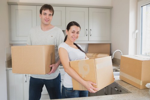 A young man and woman in a bright, modern kitchen are standing side by side, each holding cardboard moving boxes typical of house removals. The man, positioned slightly behind the woman, has short brown hair and is wearing a light grey sweater and dark trousers, carrying a medium-sized box. The woman, standing in front, has long dark hair tied back and is dressed in a white top with short sleeves and jeans, smiling while holding a slightly larger box. The kitchen features pale grey cabinetry with a smooth finish, stainless steel handles, and a white countertop with a stainless steel sink visible on the right. Natural light streams in through a window on the right, illuminating the scene. Several additional cardboard boxes are placed on the countertop and near the window, some sealed with packing tape, indicating an ongoing move. The scene captures a moment of packing or unpacking during a house removal or relocation, with the background providing a sense of a typical residential kitchen environment in Richmond, suitable for local removals services by Richmond Man with Van.