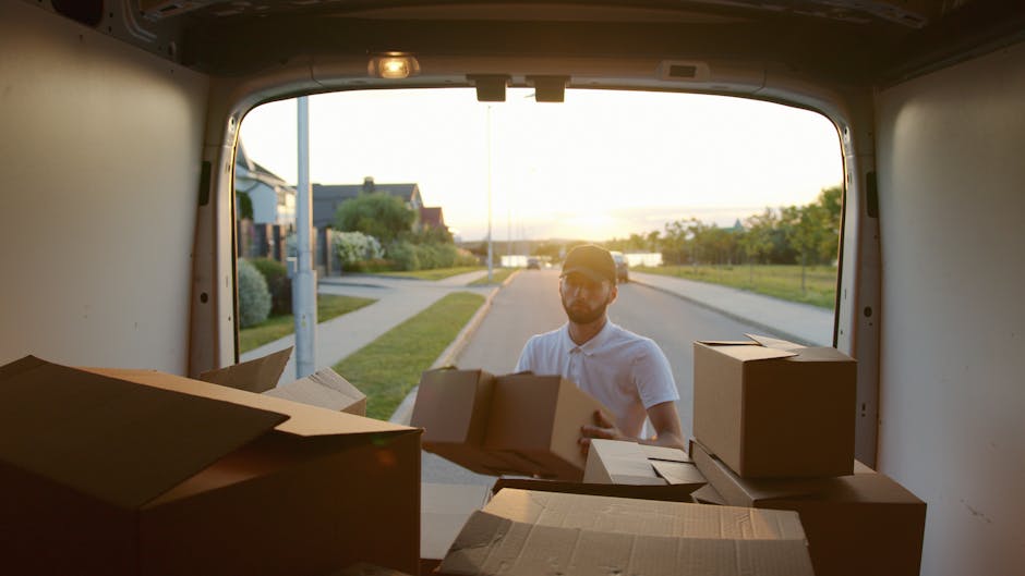 A man wearing a dark vest, black pants, and a cap is standing at the open back of a large white van parked outdoors on a paved surface, surrounded by several packed cardboard boxes of various sizes wi
