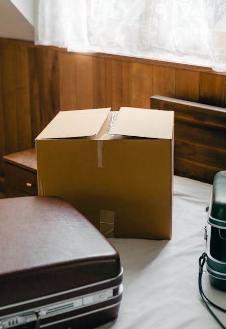 A cardboard moving box, partially open with its flaps raised, is placed on a bed in a room with wooden wall paneling and a white curtain. Next to the box, there is a closed brown leather suitcase on t